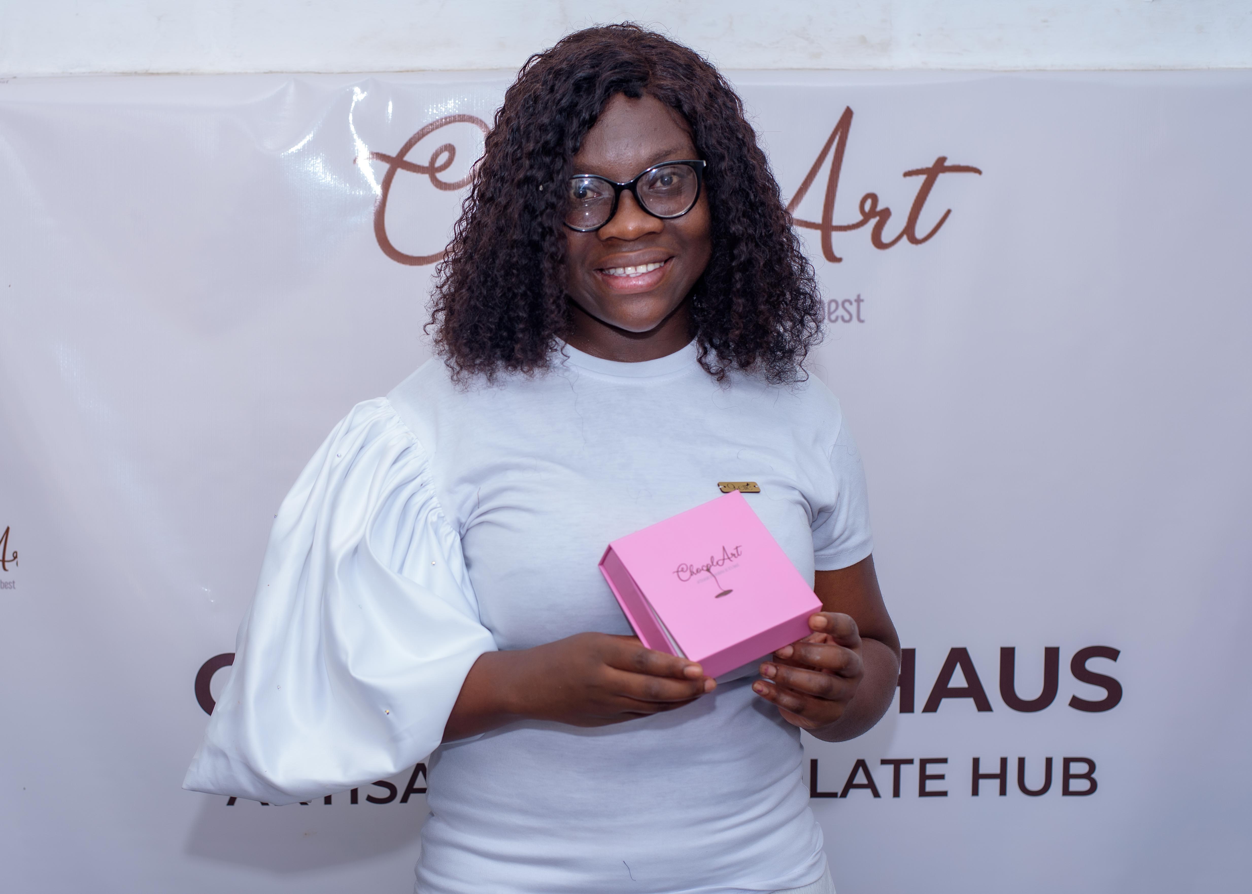 Pearl Kitcher, founder of ChocolArt Haus in Ghana, holding a pink box of handcrafted chocolate in front of her business banner.