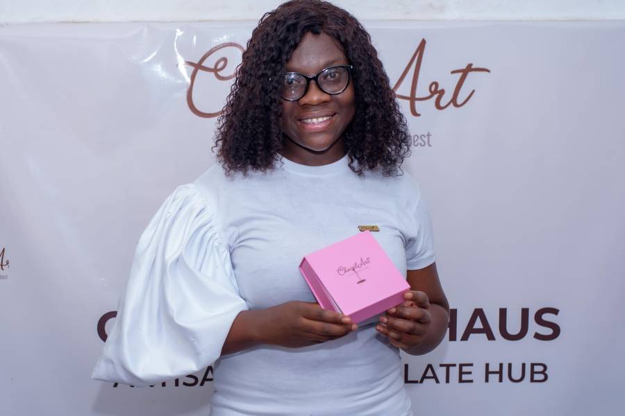 Pearl Kitcher, founder of ChocolArt Haus in Ghana, holding a pink box of handcrafted chocolate in front of her business banner.