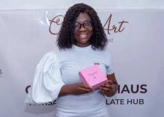 Pearl Kitcher, founder of ChocolArt Haus in Ghana, holding a pink box of handcrafted chocolate in front of her business banner.